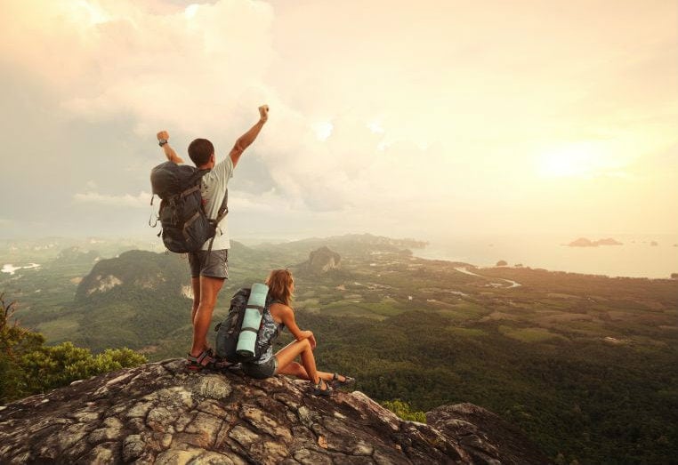 Pessoa com mochila sobre um mirante rochoso, diante de uma paisagem ao pôr do sol