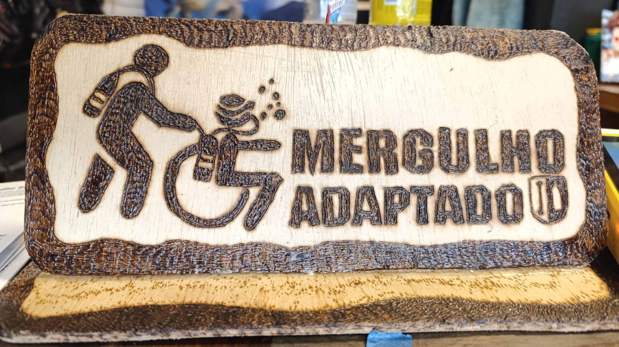 Placa de madeira entalhada com o desenho de uma pessoa em cadeira de rodas e o texto Mergulho Adaptado
